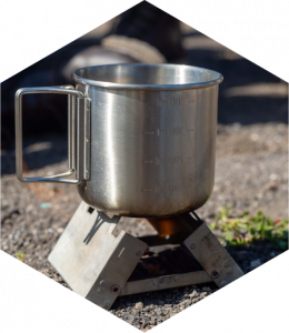 A close-up image of a metal mug over a portable camping stove. The background shows blurred dirt, suggesting an outdoor setting.