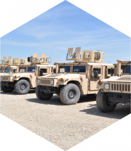A row of beige-colored Humvees parked on a gravel surface under a clear blue sky. The vehicles are equipped with various attachments and equipment on their roofs.