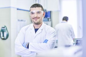 Bakelite employee Federico Rosi poses for a photo in a lab while wearing a white lab coat and safety googles. A coworker works in the background but is blurry.