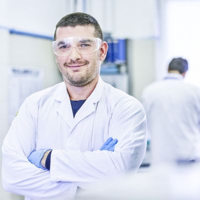 Bakelite employee Federico Rosi poses for a photo in a lab while wearing a white lab coat and safety googles. A coworker works in the background but is blurry.