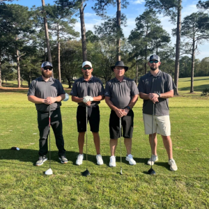 Four Bakelite employees stand on a golf course during a charity golf tournament. Trees and blue sky in the background.