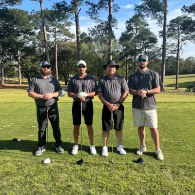 Four Bakelite Employees stand on a golf course holding golf clubs. Tress in the background.