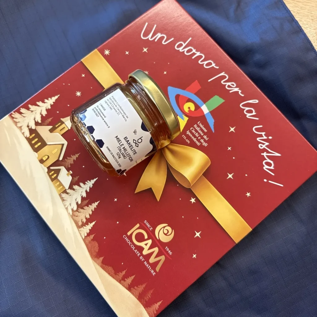 A gift set consisting of a red holiday-themed box with gold ribbon artwork and a small jar of honey placed on top, displayed on a dark blue fabric bag.