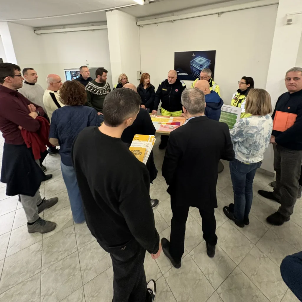 A group of people standing in a room around a table covered with safety training materials, listening to an instructor wearing emergency services attire.